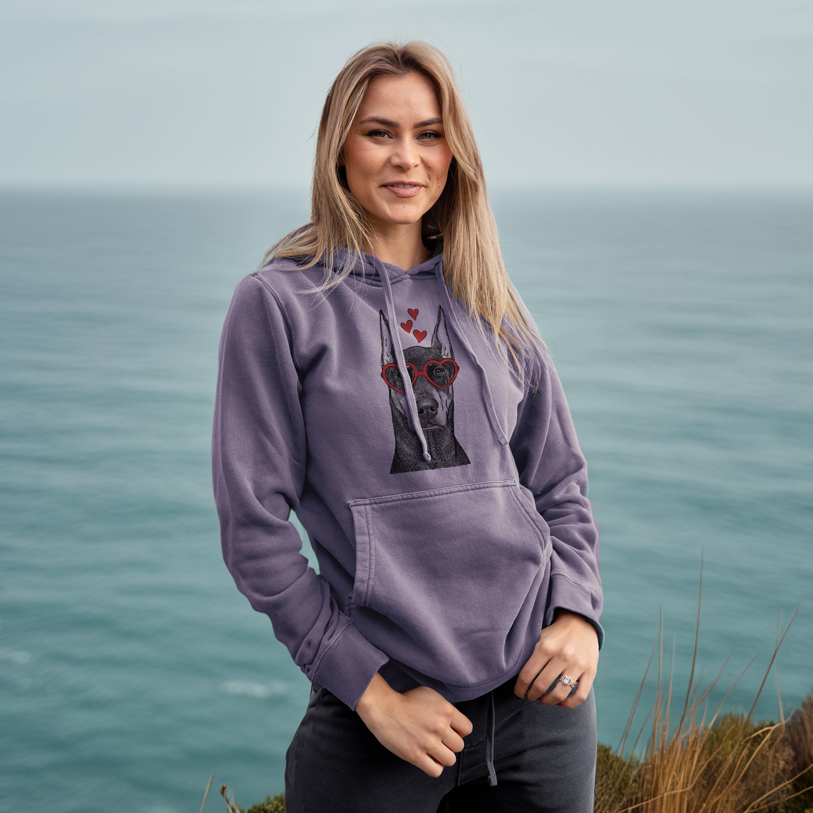 Hand-drawn sketch of Drake the Doberman Pinscher wearing heart-shaped glasses on a Plum Unisex pigment dyed hoodie, worn by a woman