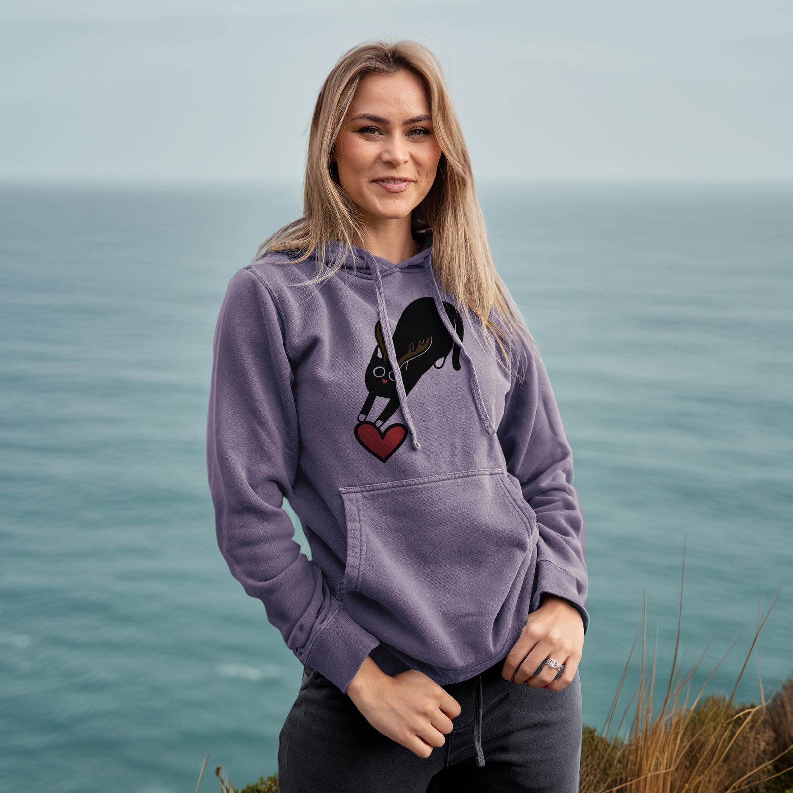 Minimalist line drawing of Bug the Black Cat with reindeer antlers and a red nose on a Plum Unisex pigment dyed hoodie, worn by a woman