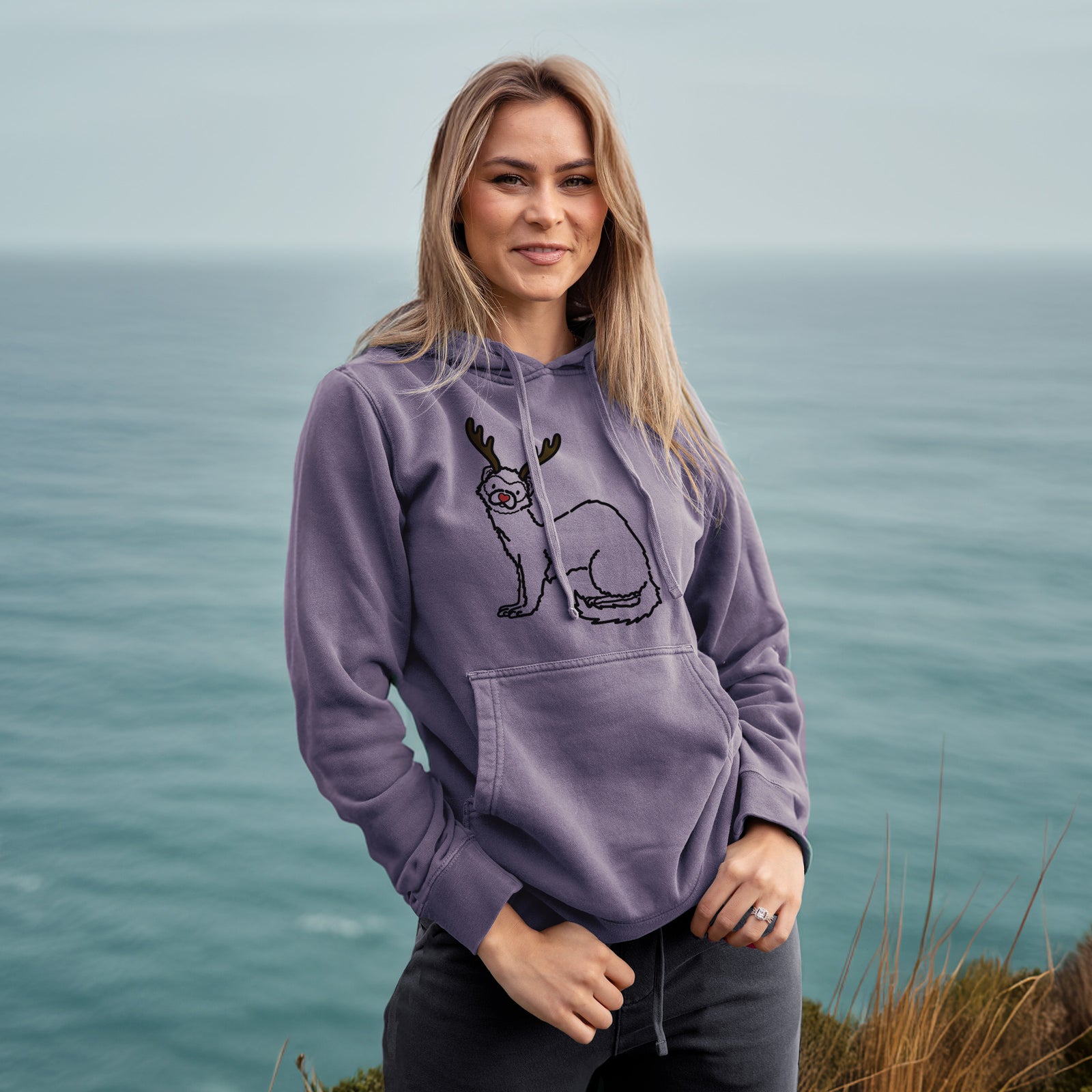 Minimalist line drawing of Fern the Ferret with reindeer antlers and a red nose on a Plum Unisex pigment dyed hoodie, worn by a woman