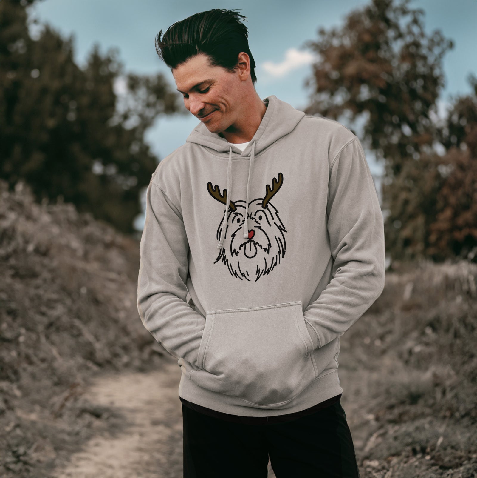 Minimalist line drawing of Tucker the Bearded Collie with reindeer antlers and a red nose on a Cement Unisex pigment dyed hoodie, worn by a man