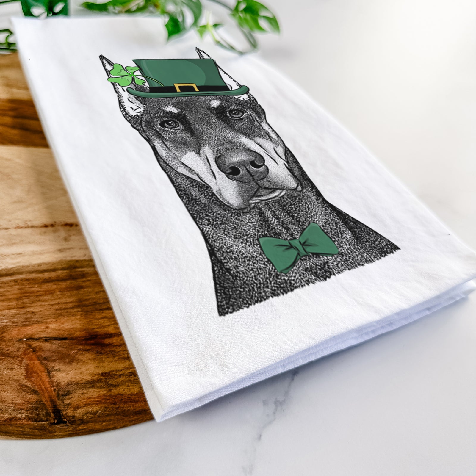 Hand-drawn sketch of Drake the Doberman Pinscher wearing a St. Patrick's Day leprechaun hat and bow tie on a tea towel