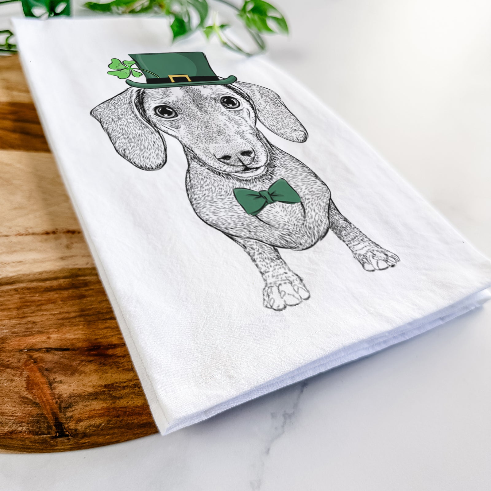 Hand-drawn sketch of Ernie the Mini Dachshund wearing a St. Patrick's Day leprechaun hat and bow tie on a tea towel
