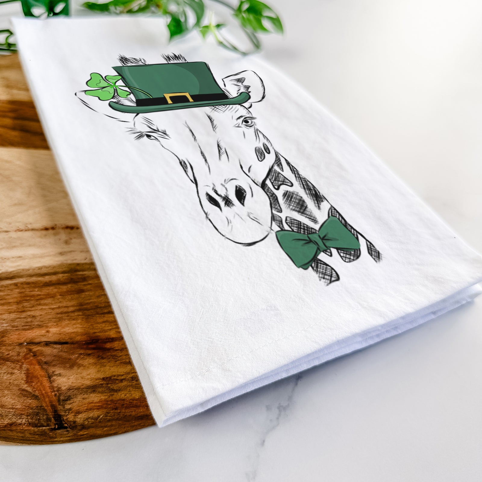 Hand-drawn sketch of Geoffrey the Giraffe wearing a St. Patrick's Day leprechaun hat and bow tie on a tea towel
