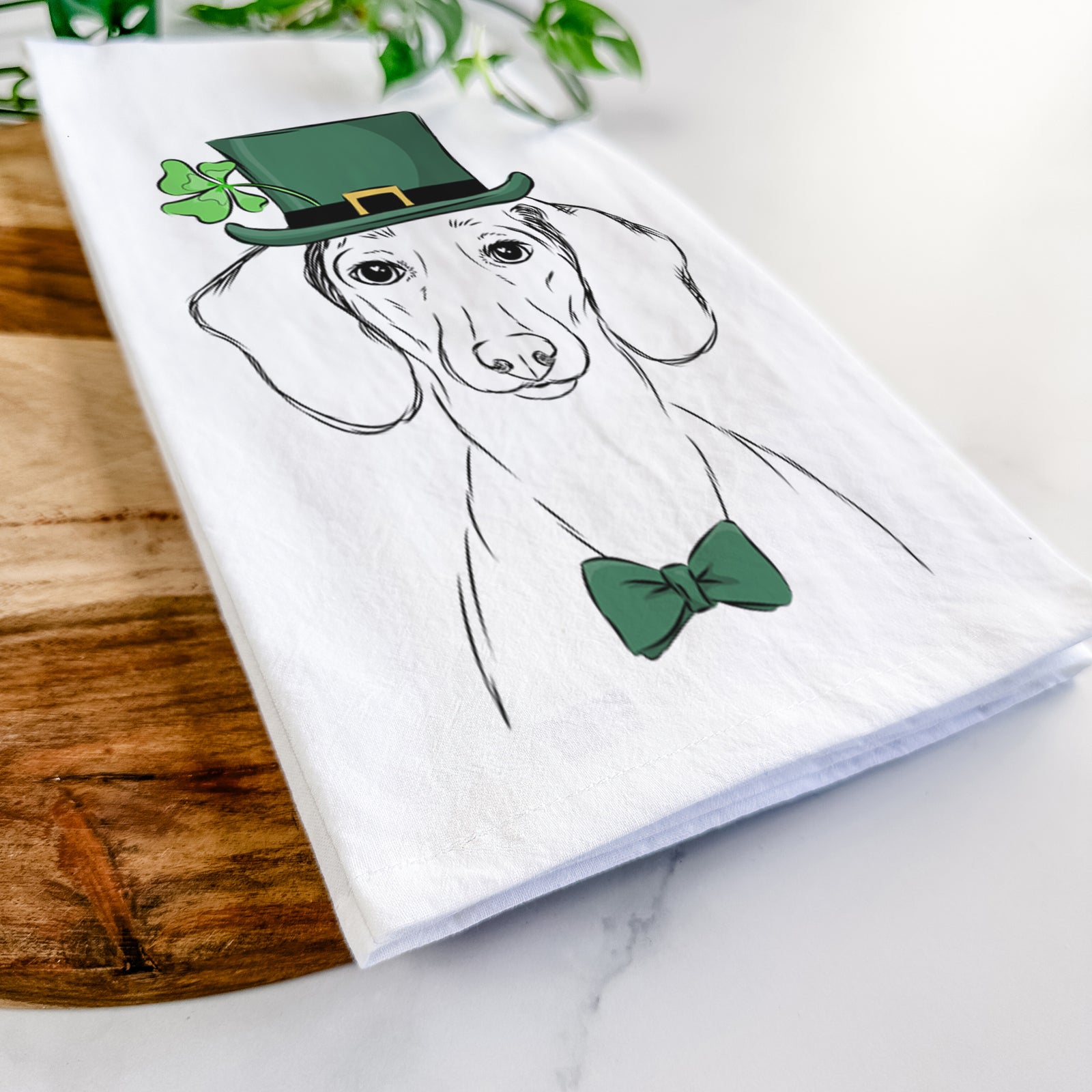 Hand-drawn sketch of Hans the Dachshund wearing a St. Patrick's Day leprechaun hat and bow tie on a tea towel
