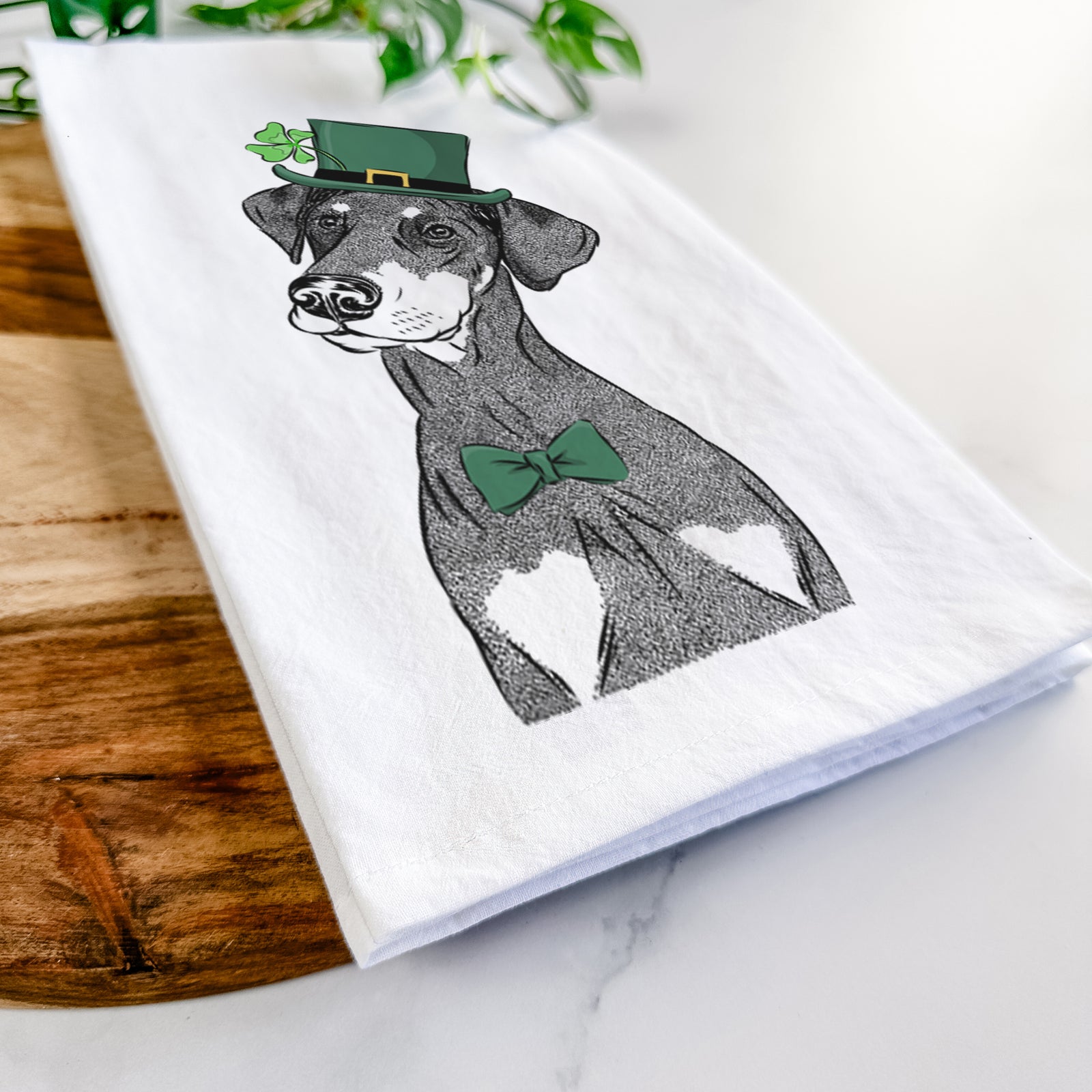 Hand-drawn sketch of Iroh the Doberman Pinscher wearing a St. Patrick's Day leprechaun hat and bow tie on a tea towel