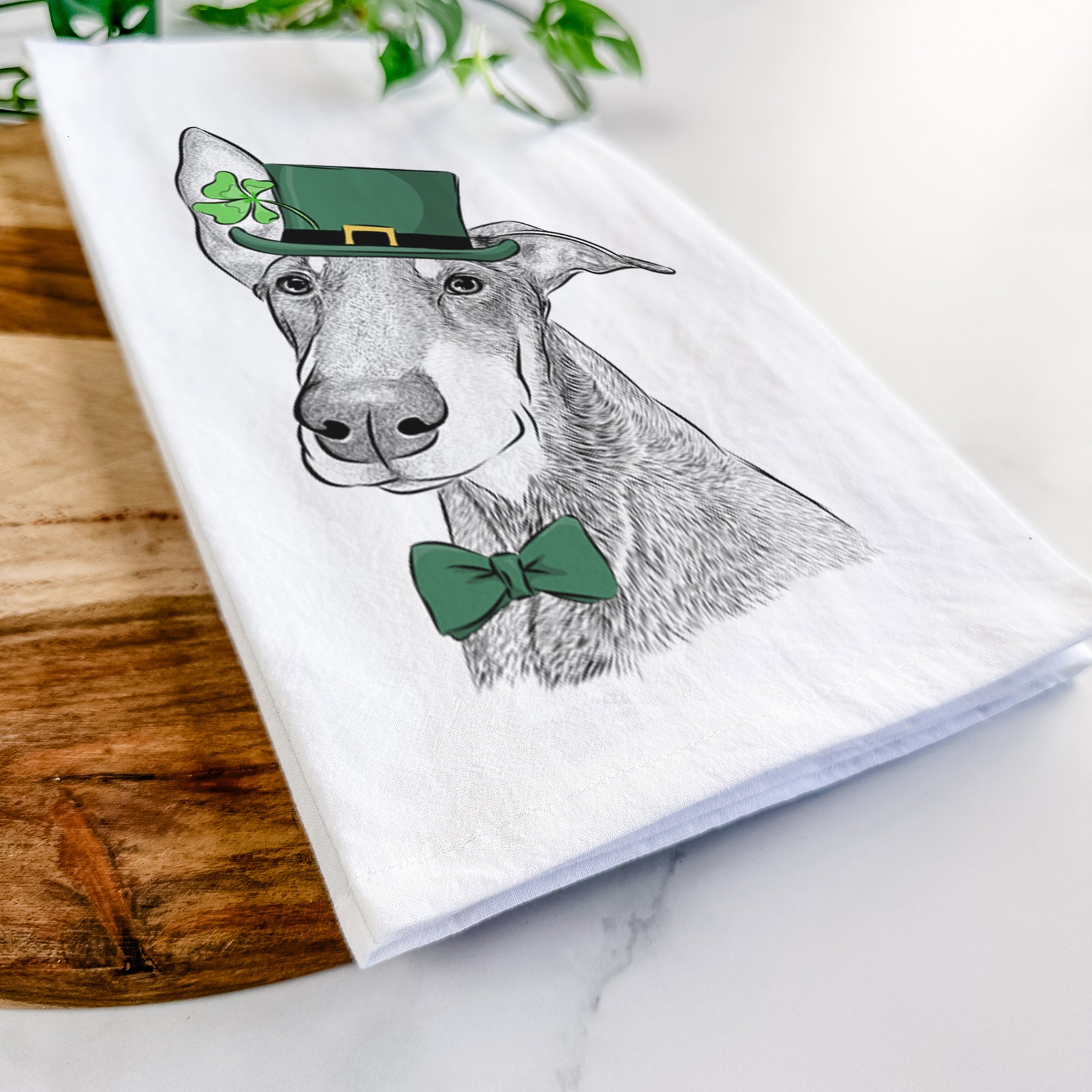 Hand-drawn sketch of Kain the Doberman Pinscher wearing a St. Patrick's Day leprechaun hat and bow tie on a tea towel