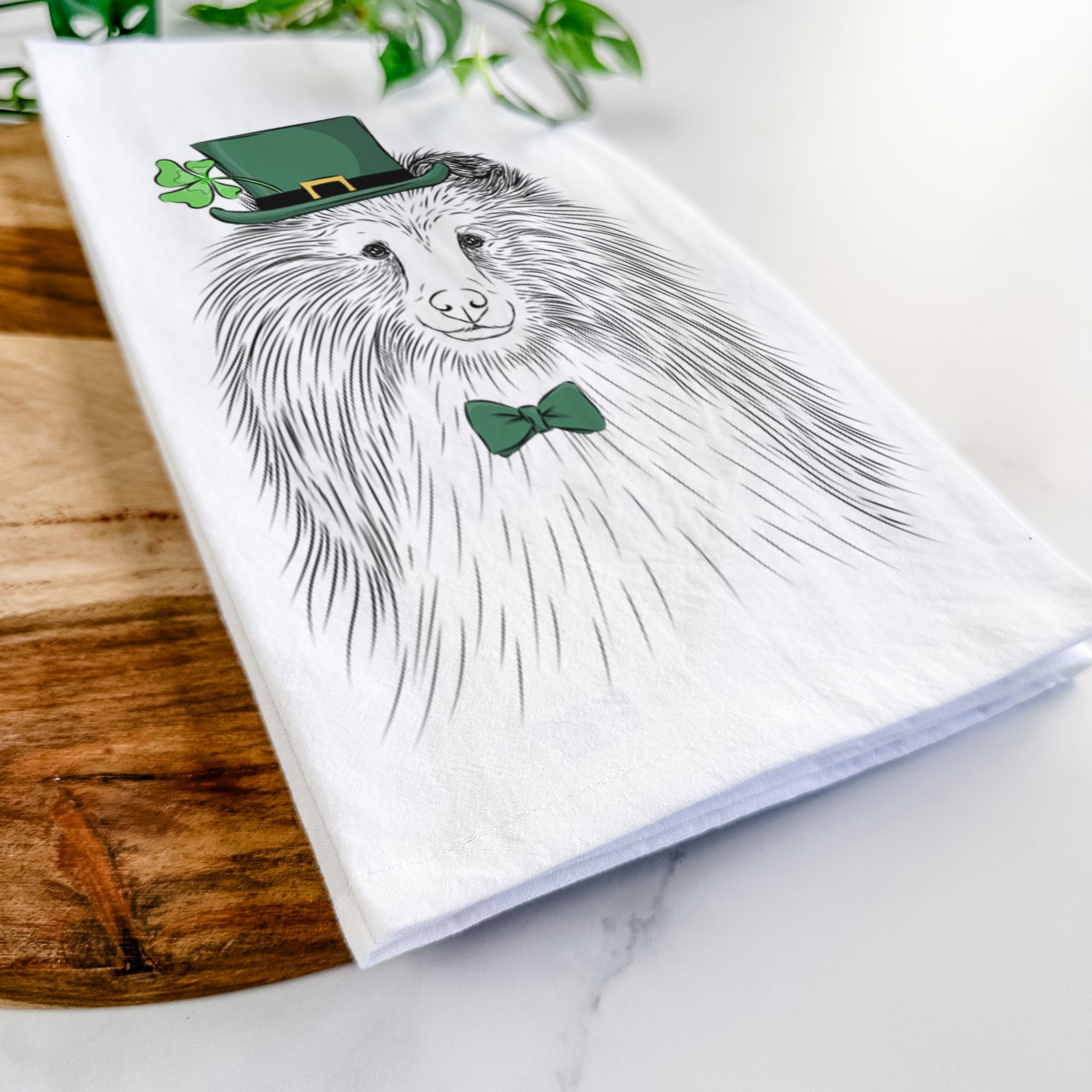 Hand-drawn sketch of Moxie the Shetland Sheepdog wearing a St. Patrick's Day leprechaun hat and bow tie on a tea towel