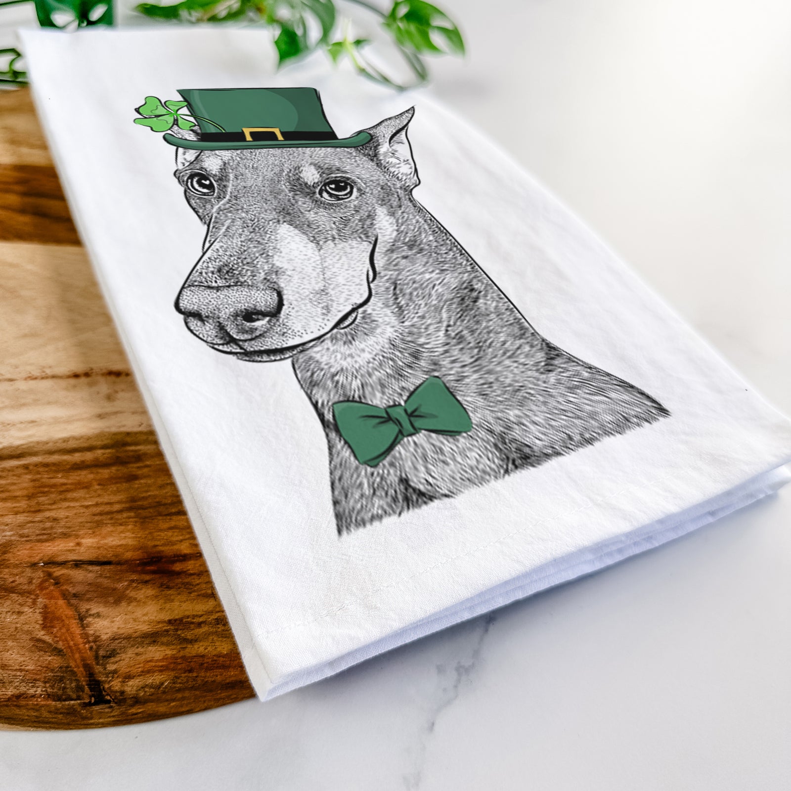 Hand-drawn sketch of Shelby the Doberman Pinscher wearing a St. Patrick's Day leprechaun hat and bow tie on a tea towel