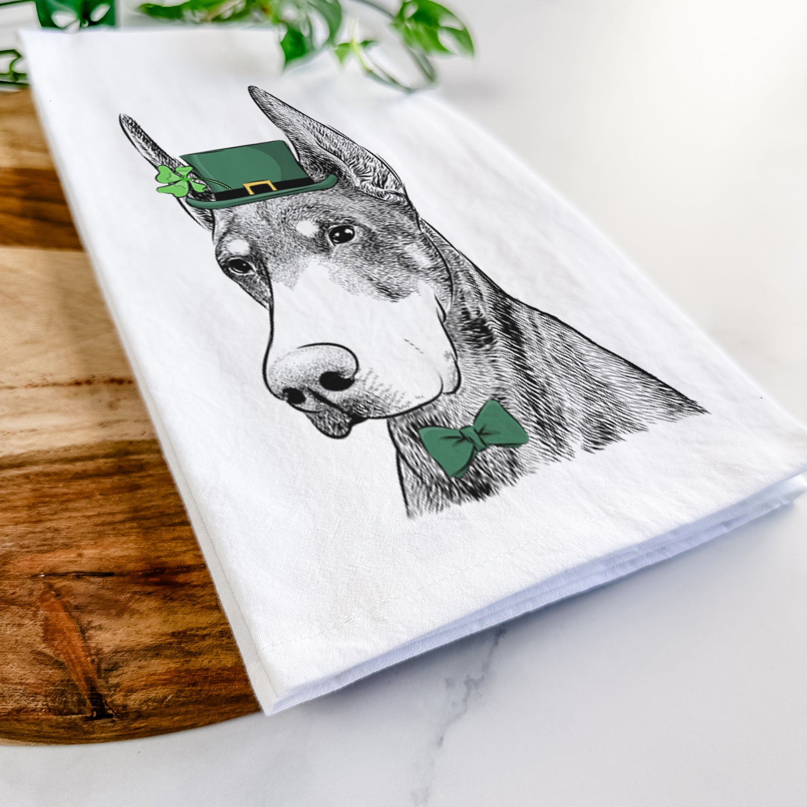 Hand-drawn sketch of Sunny the Doberman Pinscher wearing a St. Patrick's Day leprechaun hat and bow tie on a tea towel