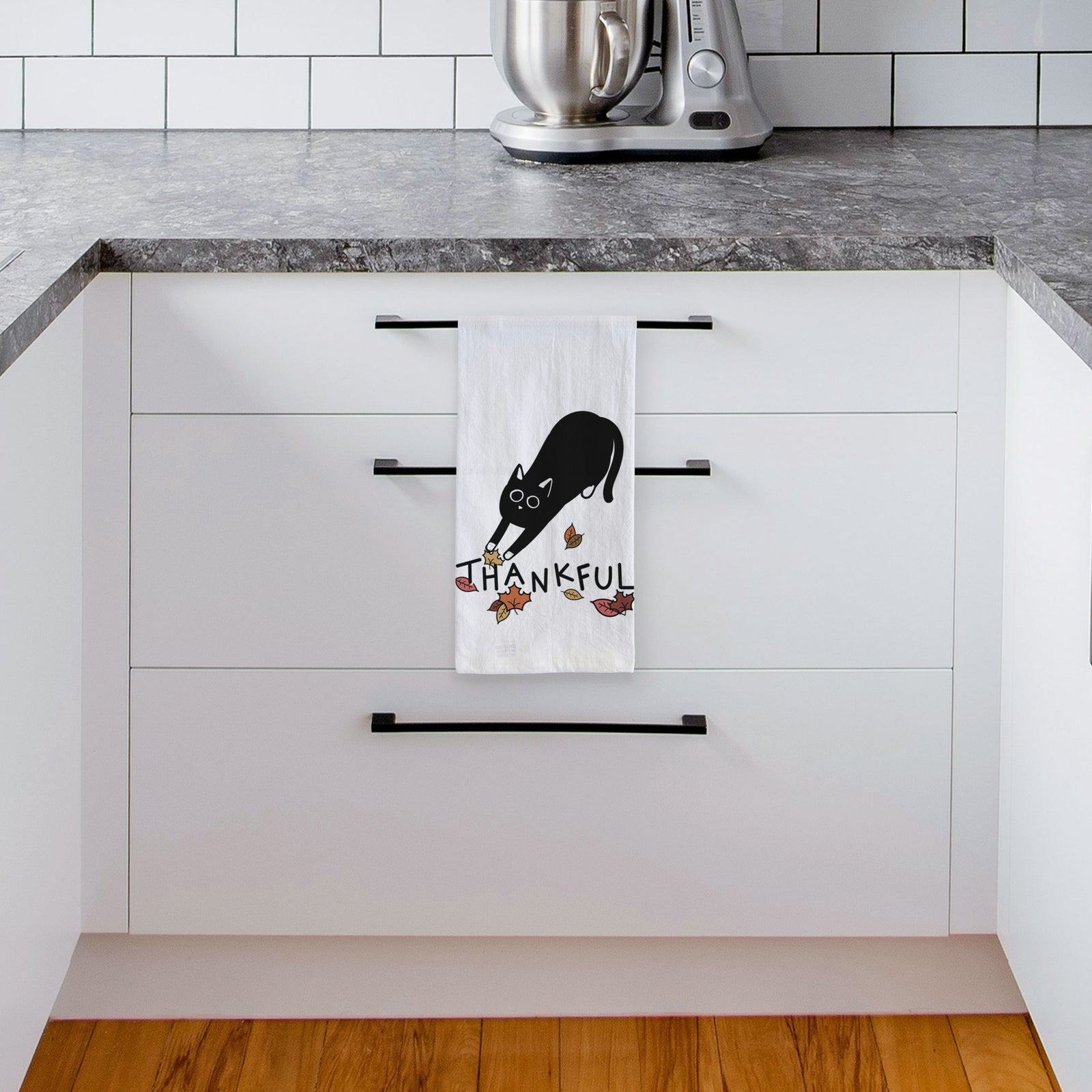 Minimalist line drawing of Bug the Black Cat with 'Thankful' lettering on a tea towel, hanging on an oven door in a kitchen