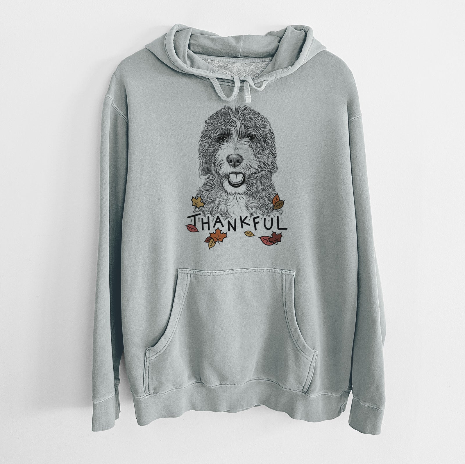 Hand-drawn sketch of Bash the Bernedoodle with 'Thankful' lettering and autumn leaves on a Sage Unisex pigment dyed hoodie
