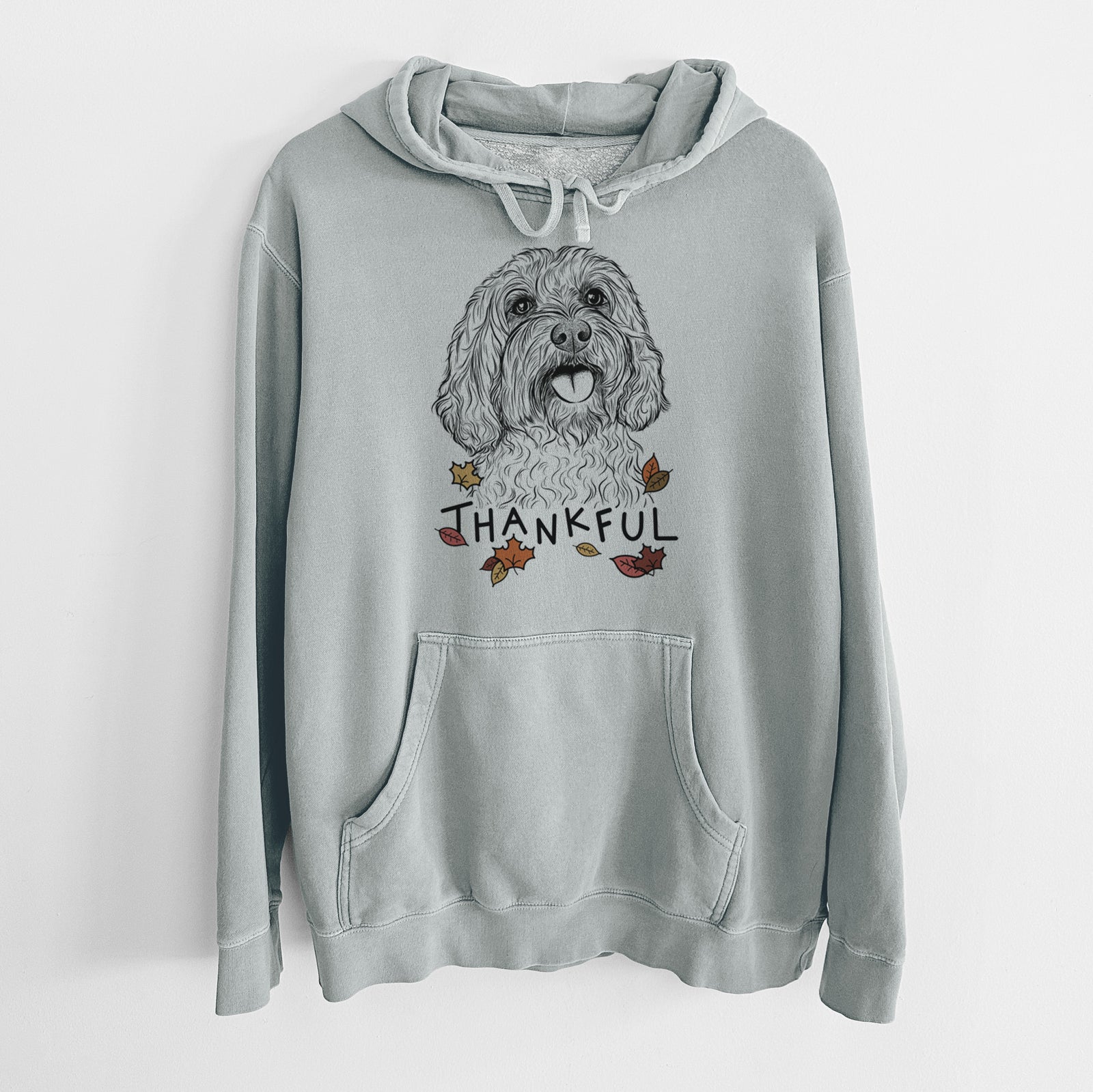 Hand-drawn sketch of Cali the Cavapoo with 'Thankful' lettering and autumn leaves on a Sage Unisex pigment dyed hoodie
