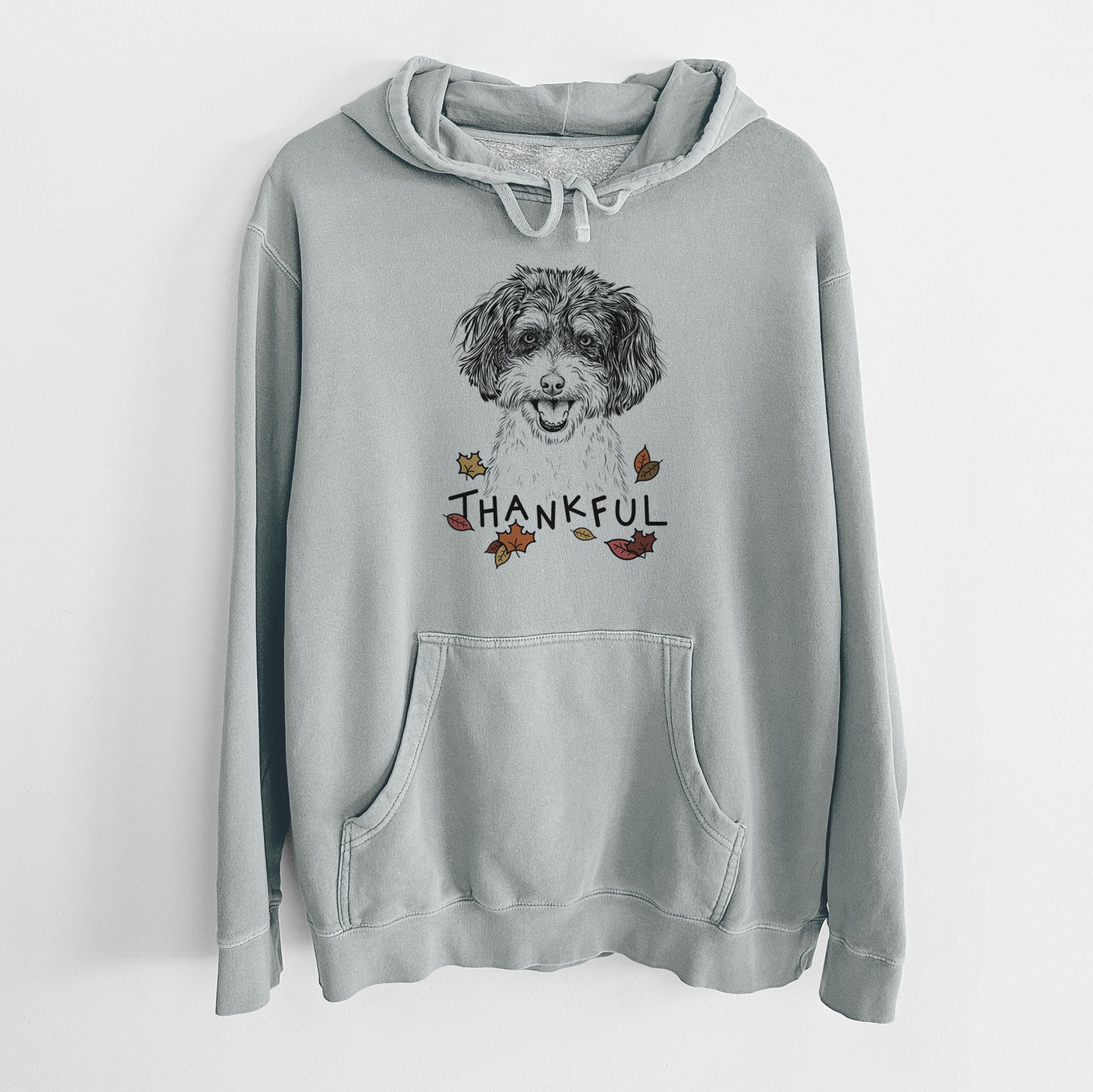 Hand-drawn sketch of Carlos the Cavapoo with 'Thankful' lettering and autumn leaves on a Sage Unisex pigment dyed hoodie