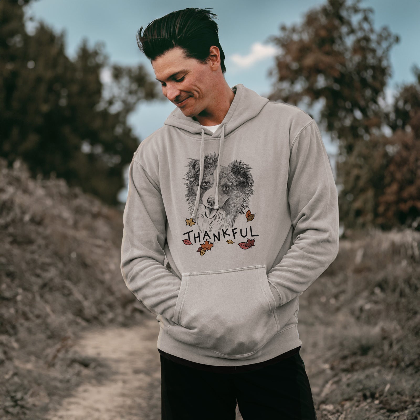 Hand-drawn sketch of Doc the Mini Aussie with 'Thankful' lettering and autumn leaves on a Cement Unisex pigment dyed hoodie, worn by a man