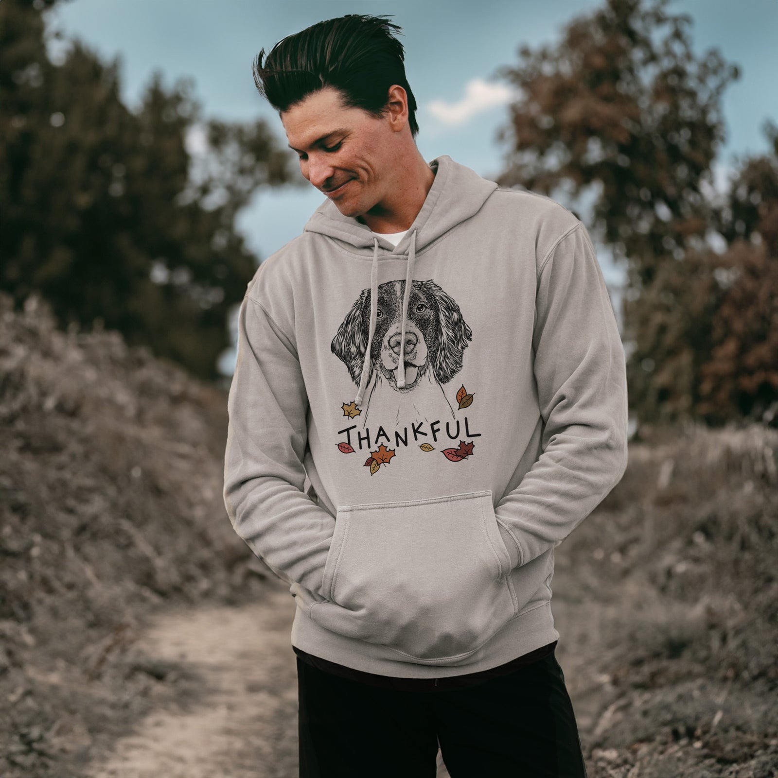 Hand-drawn sketch of Ever the English Springer Spaniel with 'Thankful' lettering and autumn leaves on a Cement Unisex pigment dyed hoodie, worn by a man