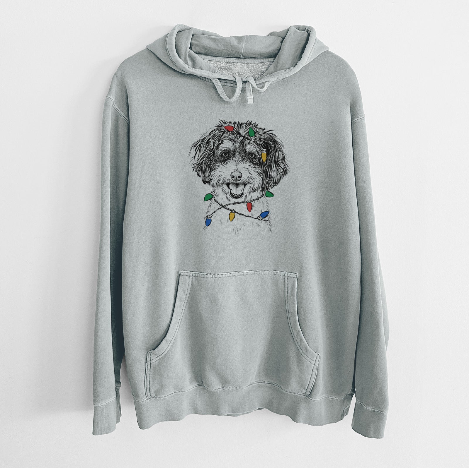 Hand-drawn sketch of Carlos the Cavapoo wrapped in Christmas lights on a Sage Unisex pigment dyed hoodie
