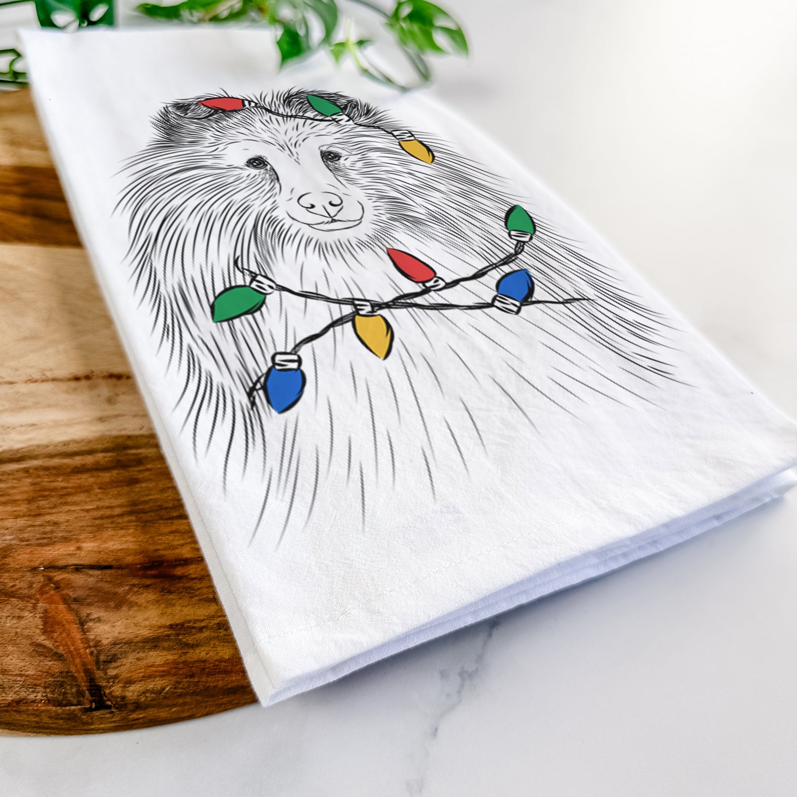 Hand-drawn sketch of Moxie the Shetland Sheepdog wrapped in Christmas lights on a tea towel