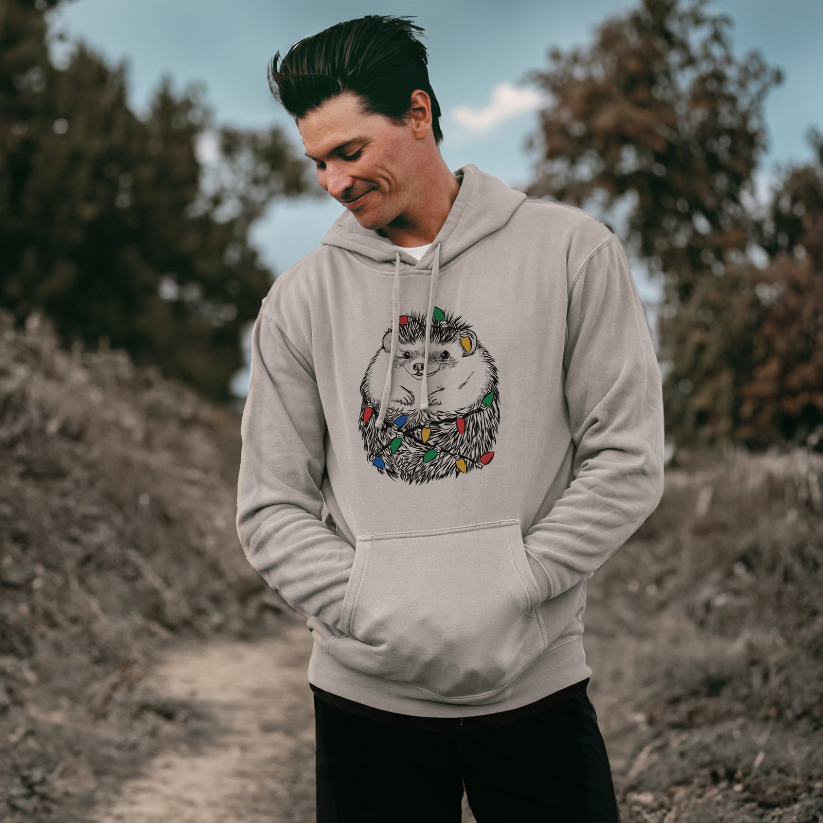 Hand-drawn sketch of Sid the Hedgehog wrapped in Christmas lights on a Cement Unisex pigment dyed hoodie, worn by a man