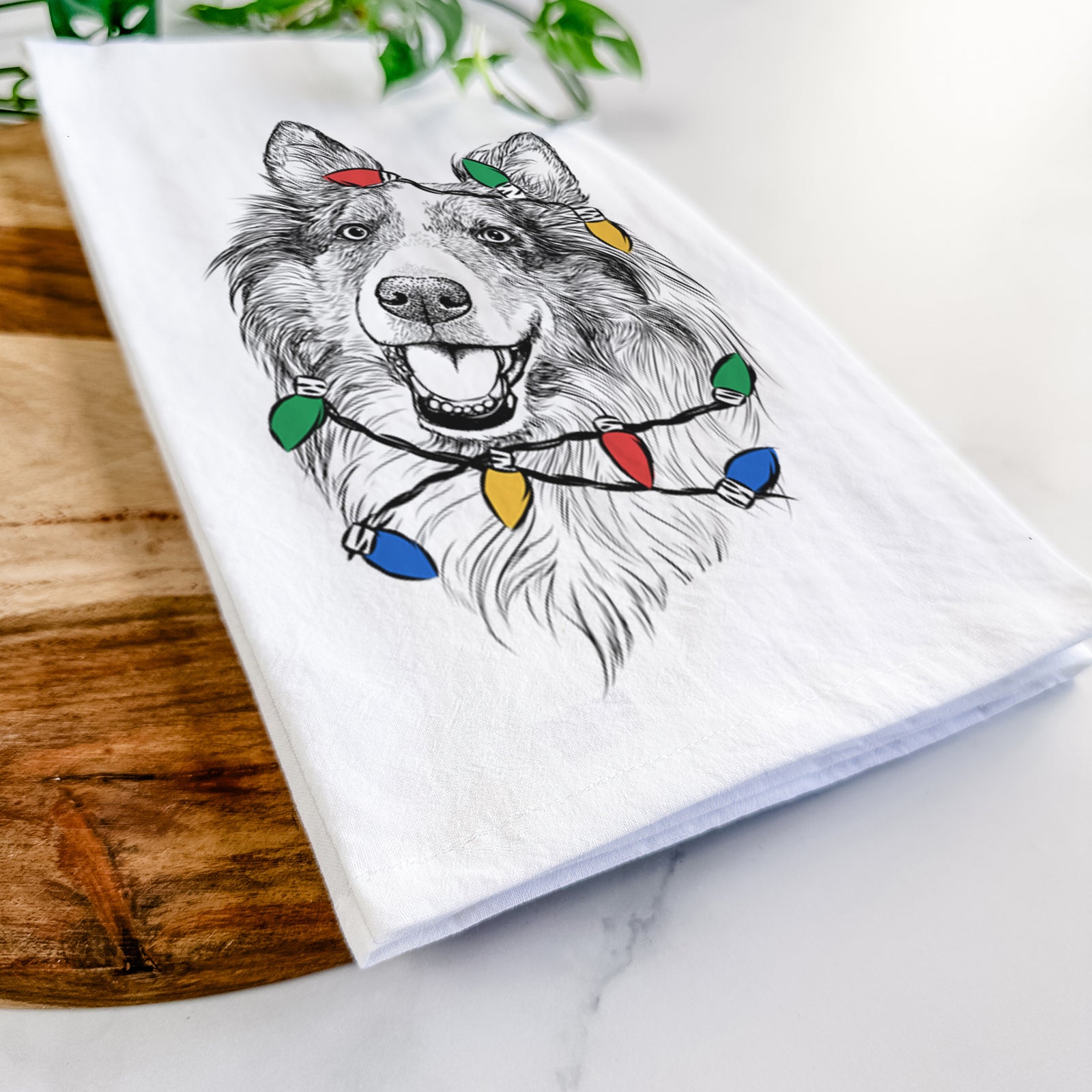 Hand-drawn sketch of Skylar the Shetland Sheepdog wrapped in Christmas lights on a tea towel