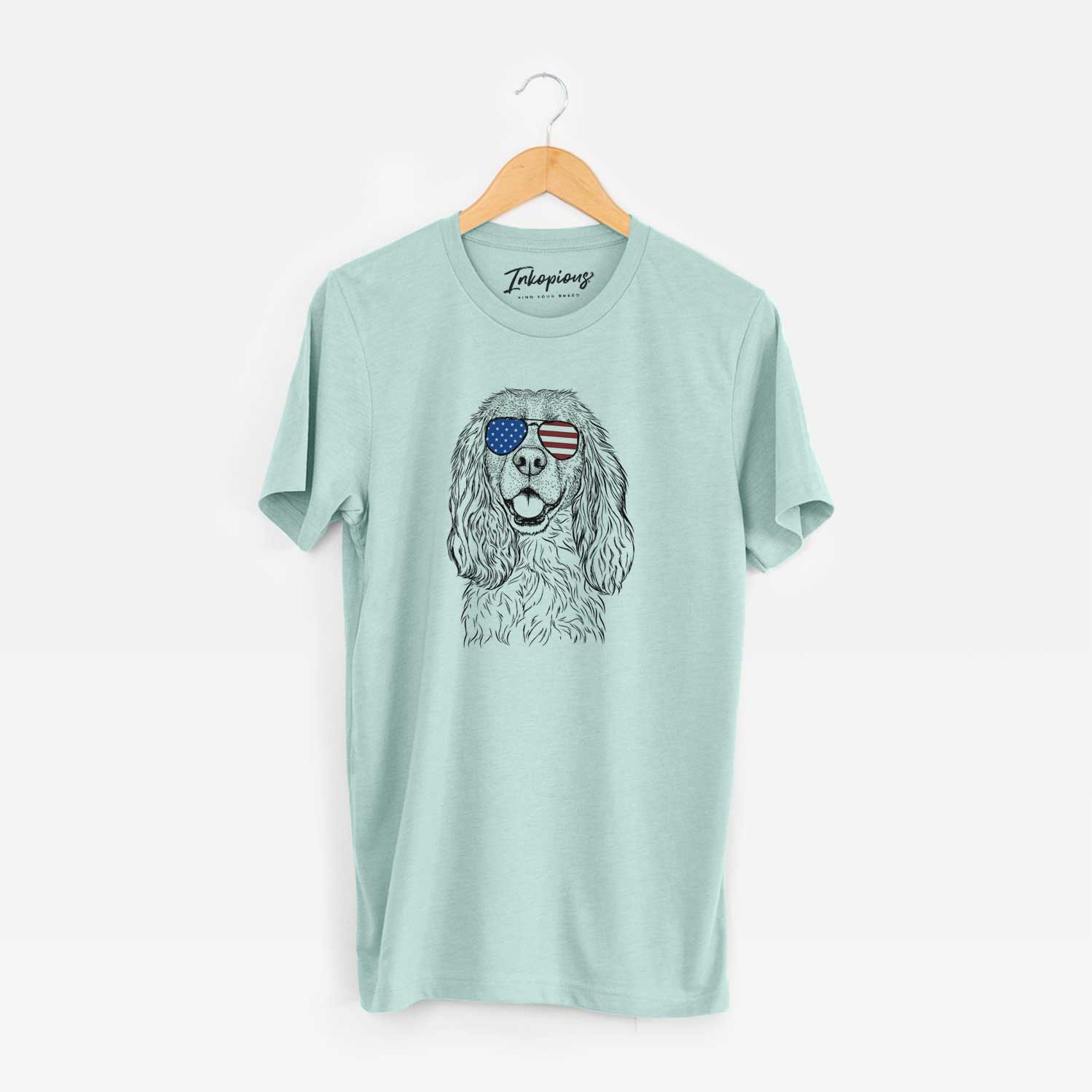 Hand-drawn sketch of Perry the Cockalier wearing American flag sunglasses on a Heather Sage Unisex crewneck