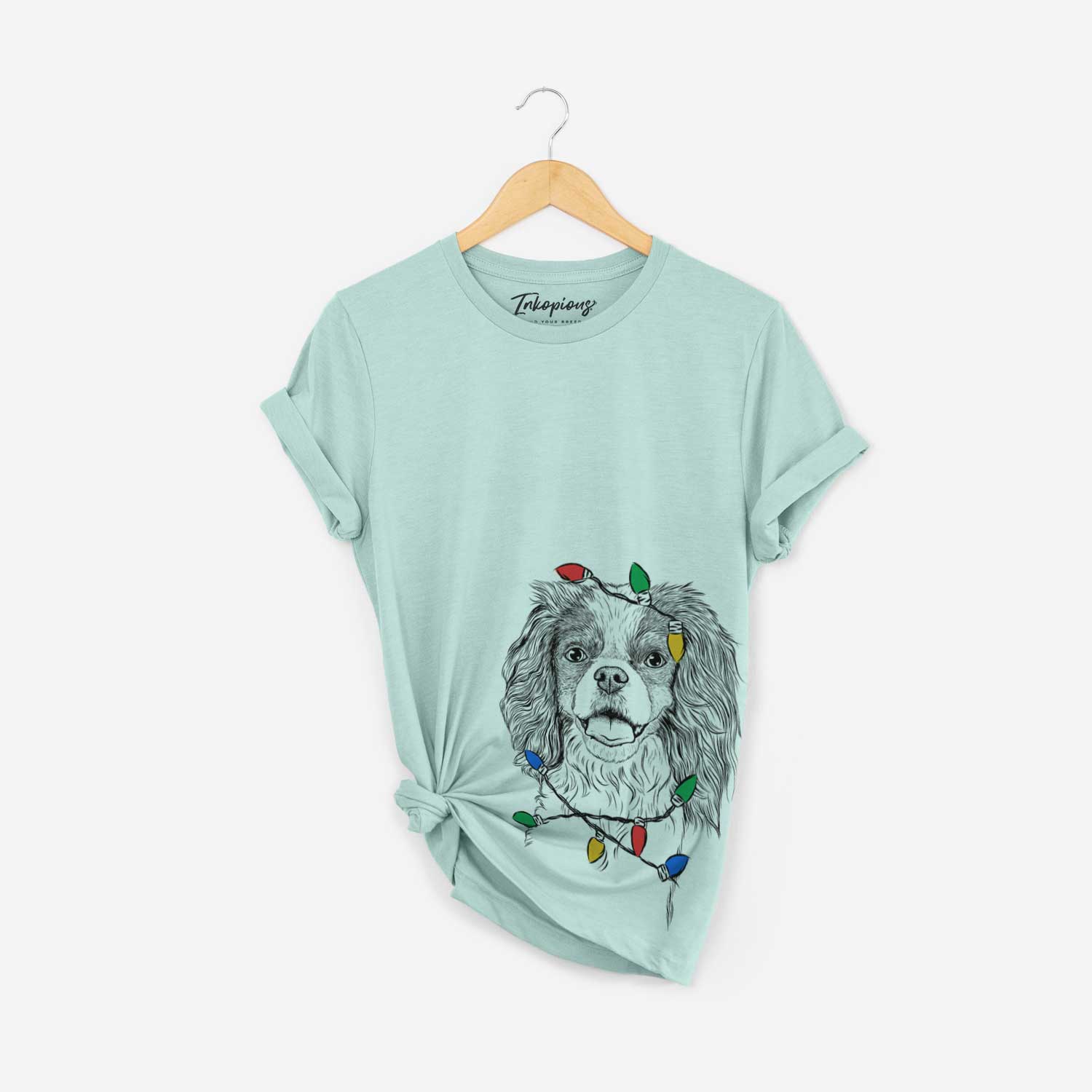 Hand-drawn sketch of Poppy the Cavalier King Charles Spaniel wrapped in Christmas lights on a Heather Sage Unisex crewneck